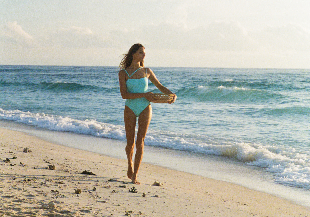 woman walking on a beach wearing a aqua blue Zabel one piece swimsuit from sustainable swimwear brand Koraru