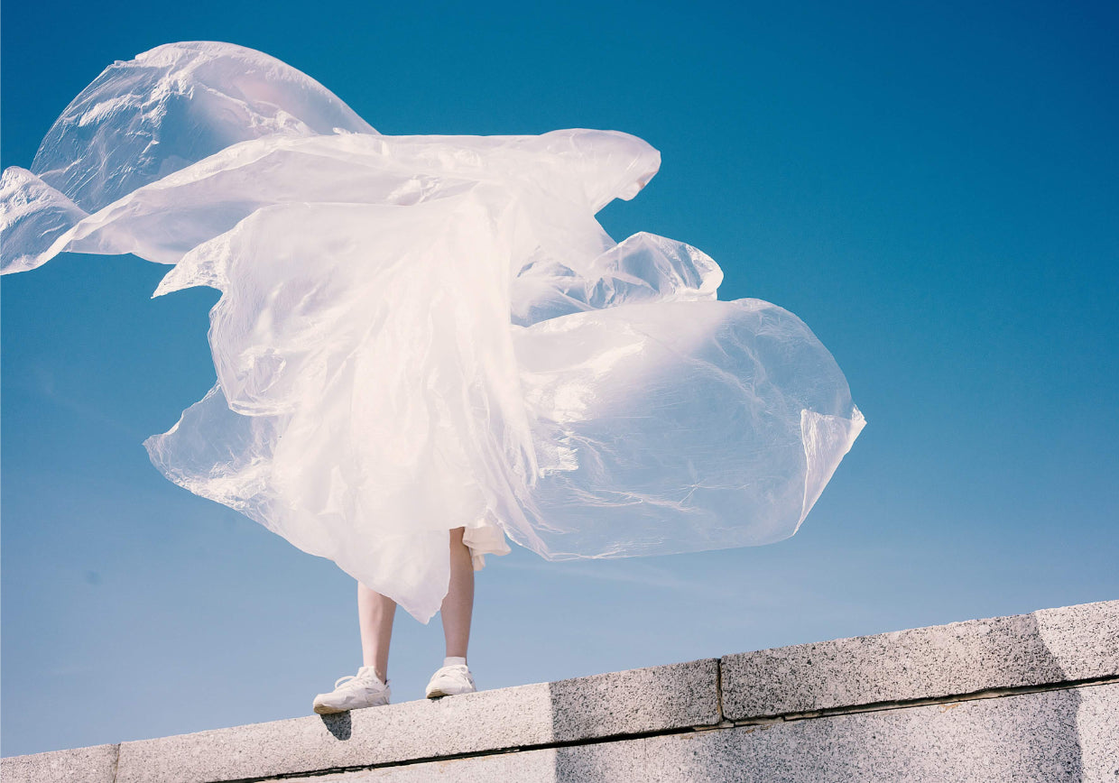 Image by Karina Tess- standing on a wall in a cloud of plastic- representing our being wrapped up in the plastic and fast fashion industry